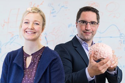 A female employee and a male employee are standing in front of a whiteboard. The female employee is smiling. The male employee smiles slightly and holds a model of a brain in his hands.
