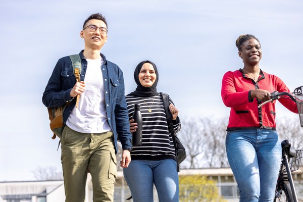 Drei junge internationale Kolleginnen und Kollegen gehen gemeinsam über den Jülicher Campus. Er hat einen Rucksack über der Schulter und eine der Frauen schiebt ein Fahrrad.