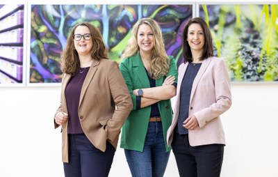 Three women stand next to each other and smile at the camera. Colorful pictures hang on a wall in the background.