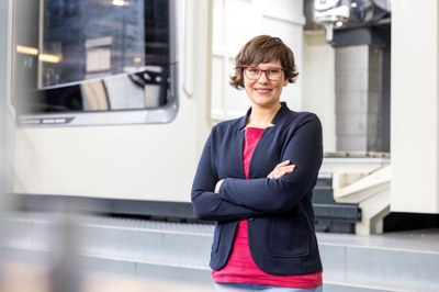 Portrait of a younger female employee in modern business attire, smiling enthusiastically into the camera, against the background of a large machine in the workshop infrastructure