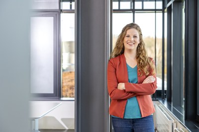 Eine Mitarbeiterin im Blazer lehnt an der Säule eines Konferenzraumes und blickt lächelnd in die Kamera.