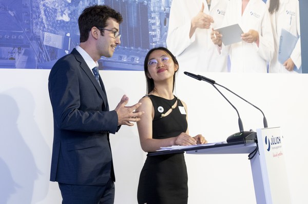 Samuel and Xinyu giving a speech at the JuDocS Ceremony (Doctoral Celebration). Xinyu is smiling at Samuel while he is talking. They are standing at a speaker's podium.