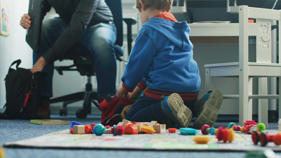 A boy sits on a carpet with his back to the camera. He is playing with toys scattered on the carpet. In front of him, a man sits on an office chair and reaches into a bag. He is only in the picture from the upper body down.