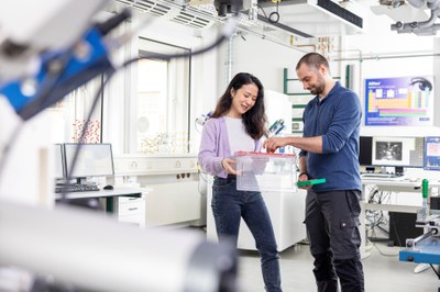 A doctoral student and a postdoc are standing in a laboratory. She has a box in her hand from which he is taking something. They both look into the box.