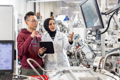 A young man in a hoodie and a young woman with a headscarf and white coat are standing in the centre of a technical system. They are looking at a digital screen.