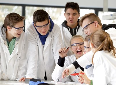 Four boys and three girls are gathered around a table, laughing. They are wearing white coats and most of them are wearing protective goggles.